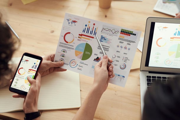 Zwei Personen analysieren bunte Diagramme auf Papier, Smartphone und Laptop.